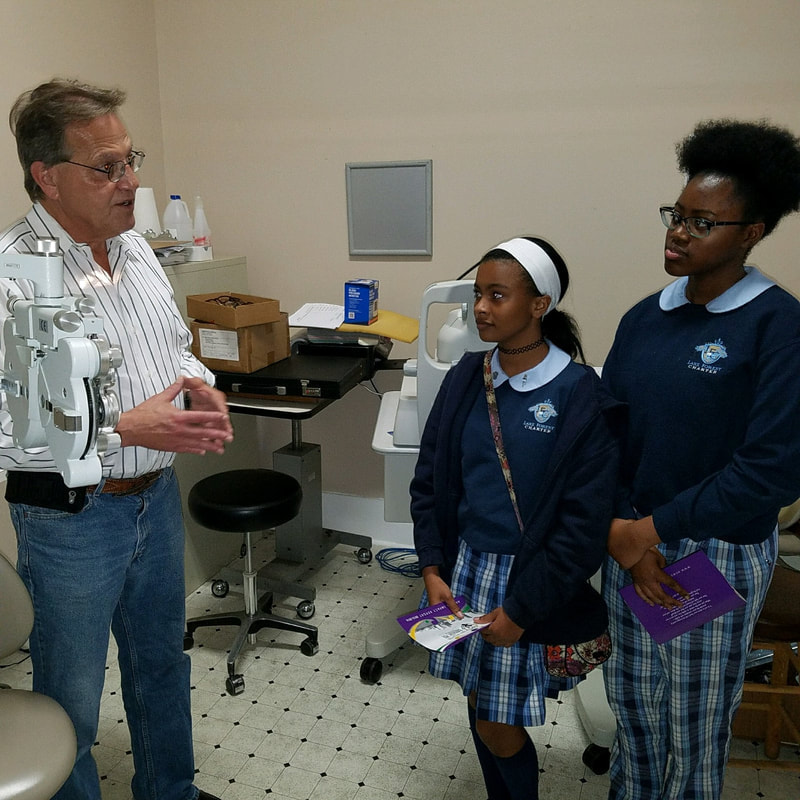 2 Black girls on the right side watch as an older white man explain the optometry equipment next to him. The first girl is shorter with medium brown skin, her hair flat-ironed and pulled back into a ponytail with a white headband, a black choker around her neck, and a long, patterned pink cross-body purse. The second girl has darker brown skin, her natural hair up in a puff, and glasses. Both girls wear matching school uniforms: a navy blue sweater with an eagle logo on the righthand side, light blue collars peaking from the neckline, and navy blue and light blue plaid bottoms. The first girl wears a skirt for her bottoms with navy blue knee-high socks. The second wears pants. Both students hold purple pamphlets in their hands. The white man on the left wears glasses, a striped button down, and jeans with a brown belt. The rest of the office is seen in the background with medical equipment, a table with a circular black chair, and an empty poster holder.