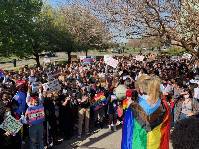 A teenager with a Progress Pride flag draped around their body holds a red and white megaphone up to their mouth in front of a crowd of roughly 100 students or more. The students hold an array of colorful signs in protest of anti-trans legislation. Trees can be seen in the background.