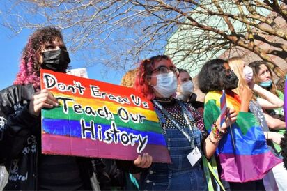 A row of students of varying ethnicities stand looking off to the right. The first student has curly brown and pink hair, dark black eye shadow, and a black leather jacket. They hold a cardboard sign painted with the pride frag colors. The words 