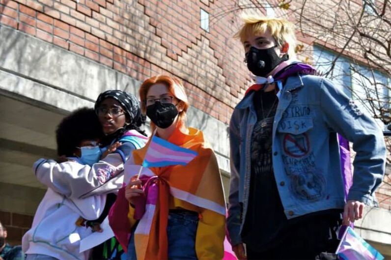 4 teenagers are seen standing solemnly in front of a crowd. The first two students are holding one another, as the one on the left cries. The crying student has a medium sized black afro and glasses. The rest of their face is obscured by a blue mask. The student holding them has dark brown skin, faux locs covered by a black bandana, and a trans pride flag wrapped around their back. Next of the 4 students is a teenager with brightly dyed orange hair, a black face mask, a lesbian pride flag wrappd around their body, and a trans pride flag in hand. The final of the 4 students has shaggy blond hair, a black face mask, a progress pride flag wrapped around their neck, a blue jean jacket, all black clothing underneath, and a smaller trans pride flag in their hand.
