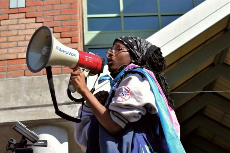A Black femme with dark brown skin is seen speaking through a red and white megaphone. Her faux locs are covered by a black bandana. They wear a trans pride flag around their body. They also wear an oversized grey hoodie and glasses.