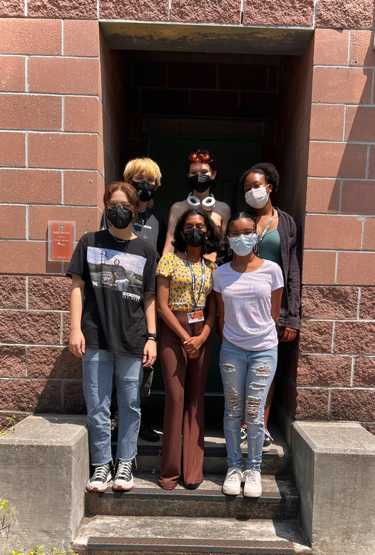 6 teenagers are seen standing in 2 rows on top of a concrete staircase. All 6 students are wearing face masks. From left to right of the back row, the first teen has shaggy bleached blonde hair with a thick black mask on and a black shirt. The second student has orange curly hair, bare shoulders, and white headphones around their neck. The third student is a little bit shorter with brown skin, a small afro, a faded teal top, and a greyish brown jacket on top. In the bottom row, there is a lightskin femme with brownish blonde hair tied into a pony tail and glasses. They wear a black t-shirt with what looks to be an album cover, jeans, and converse. The next student is much shorter with short black hair that's curcled at the end, a patterned yellow top, an orange student ID, and long brown pants. The final student also has brown skin, her black hair slicked back into a ponytail with glasses, hoop earrings, a white t-shirt, ripped pastel jeans, and white Nike tennis shoes.
