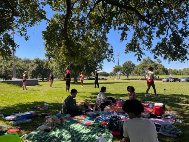 Several teenagers of varying ethnicites are seen sitting on various patterned picnic blankets under the shade of a large tree. In the background, other teenagers can be seen tossing a volleyball back and forth in the sunlight, standing in the middle of a grassy field. Other trees can be seen in the distance as well as parked cars and a bright, blue sky.