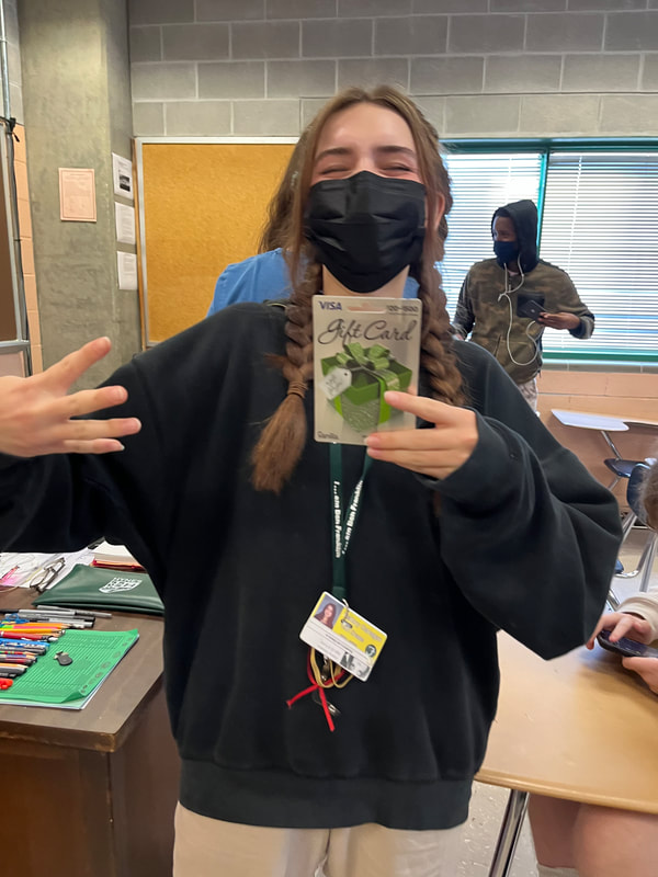A white student with long blond hair in two braids smiles underneath her black face mask, showing her VISA gift card proudly. She is dressed in a black hoodie with a yellow student ID. She is seen in a classroom with other students and desks behind her.