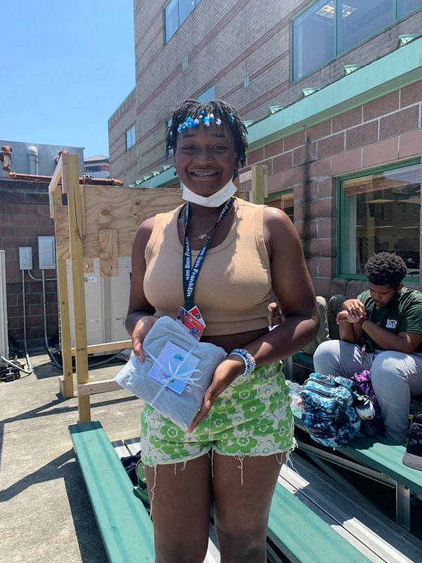 A darkskin Black student proudly showcases their carefully wrapped binder with a note from BFP on top. The student has braces, twists with light blue and white beads on the end, an orange Student ID, a tan tank top, and patterned green and white shorts. They seem to be standing on green bleachers outside with another Black student staring at his watch in the background, a blue bookbag in between his legs.