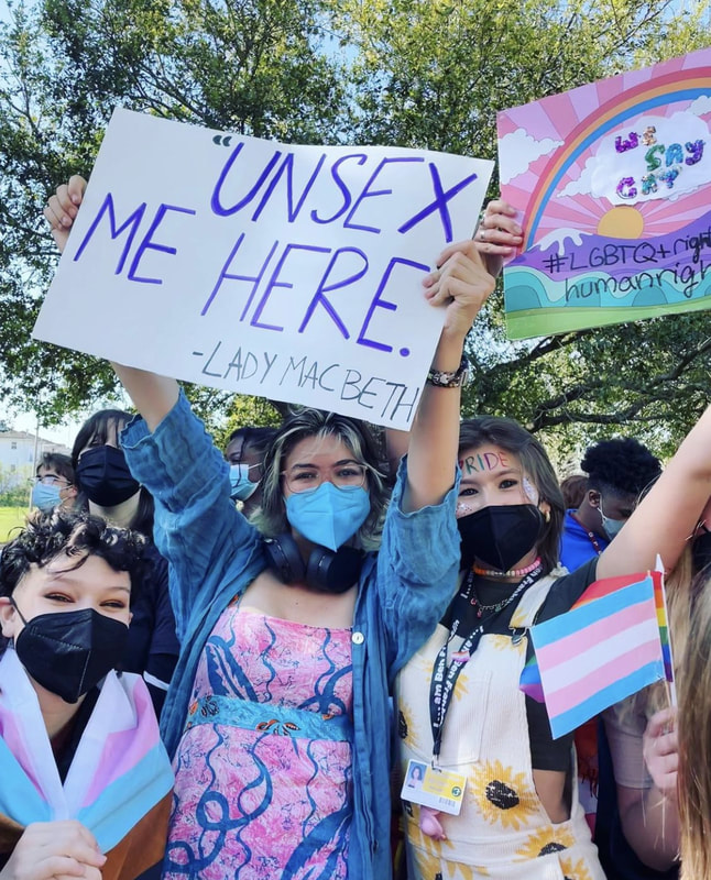 3 white teenagers stand out amongst a crowd, smiling into the camera. The first is crouched down with curly black hair, a black face mask, and a trans pride flag around their body. The second student is standing up, wispy black and bleached blonde hair and a blue face mask. She is wearing a blue jean jacket and a pink and blue patterned dress. She proudly holds up a sign that says, 