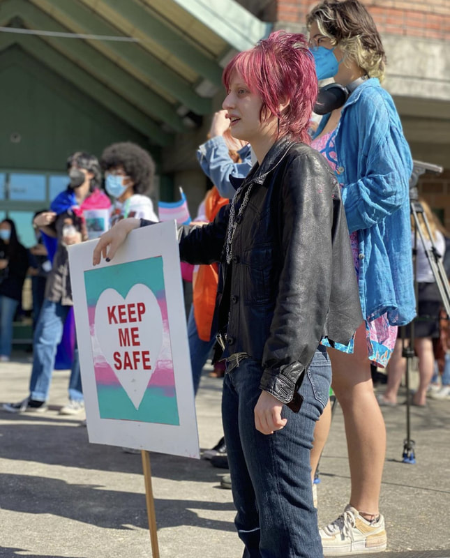 A white masc-presenting teen is seen standing in a black leather jacket and dark denim pants. Their hair is wispy and dark red. They lean their hand on a sign with a stick attached that reads 