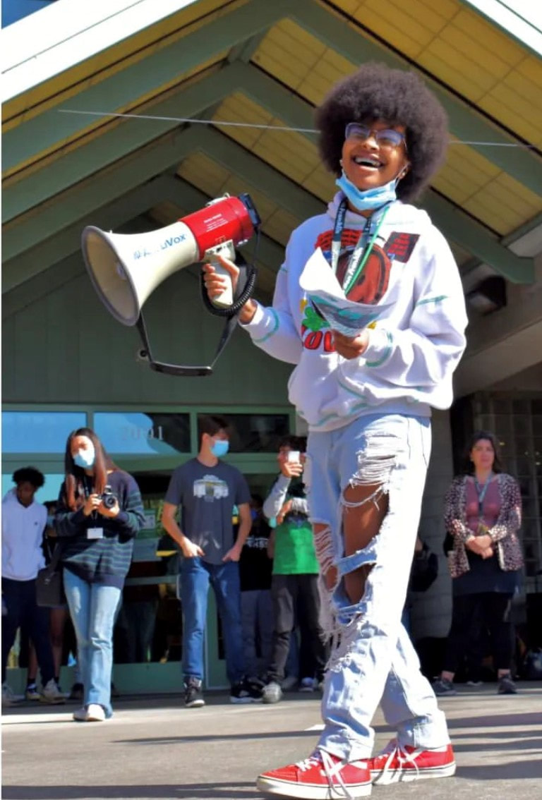A Black femme with medium brownskin laughs with their eyes closed in front of a crowd. Her hair is a medium sized afro. She wears an oversized white hoodie with a smiling brown face on it and the word 