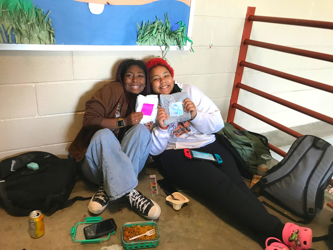 Two Black students can be seen sitting down on a concrete floor next to the red metal railing for a staircase. Their bags, phones, and food are displayed in front of them. The two students proudly display their carefully wrapped chest binders with little notes attached on top.